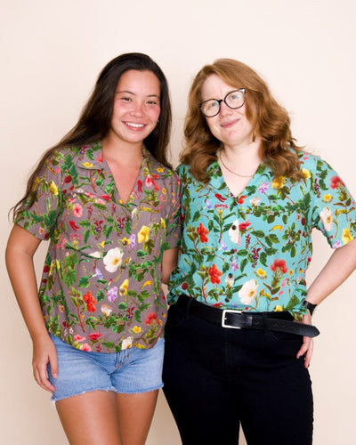 Two smiling models showcase button-up collared shirts with an intricate print of native Hawaiian plants. The model on the left has long brown hair and a poi-colored floral shirt, while the model on the right has medium-length red hair, glasses, and a kai colored floral shirt.