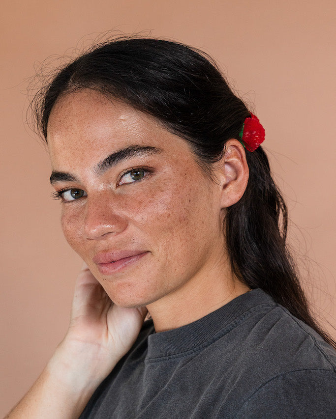 Woman with dark brown hair and red 'ohi'a lehua hair clip behind her ear smiles at the camera