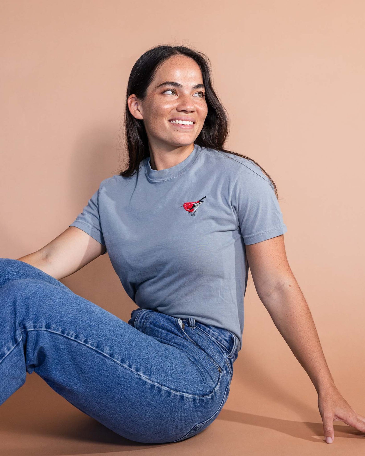 Woman wearing jeans and a light gray t-shirt with a small red bird embroidered on the top right corner