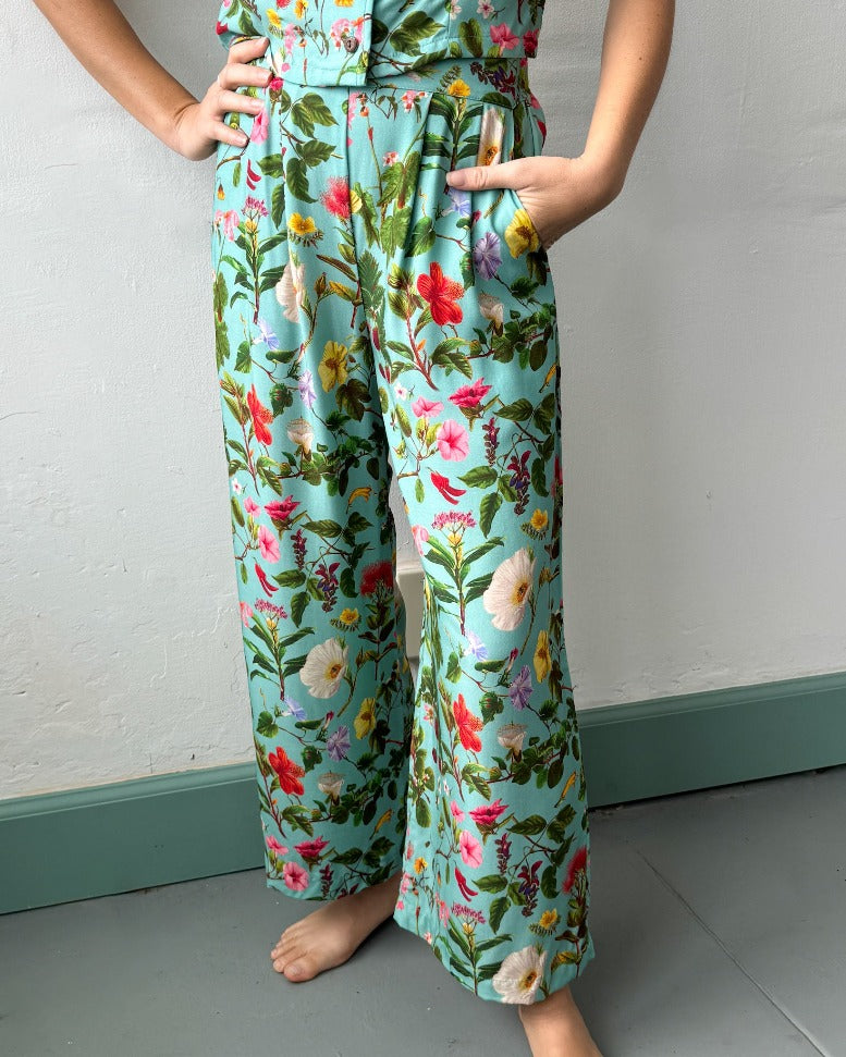 A model, visible only from the waist down, has her right hand on her hip and her left hand in her pocket. She's wearing vibrant blue pants with a complex pattern of native Hawaiian flowers.