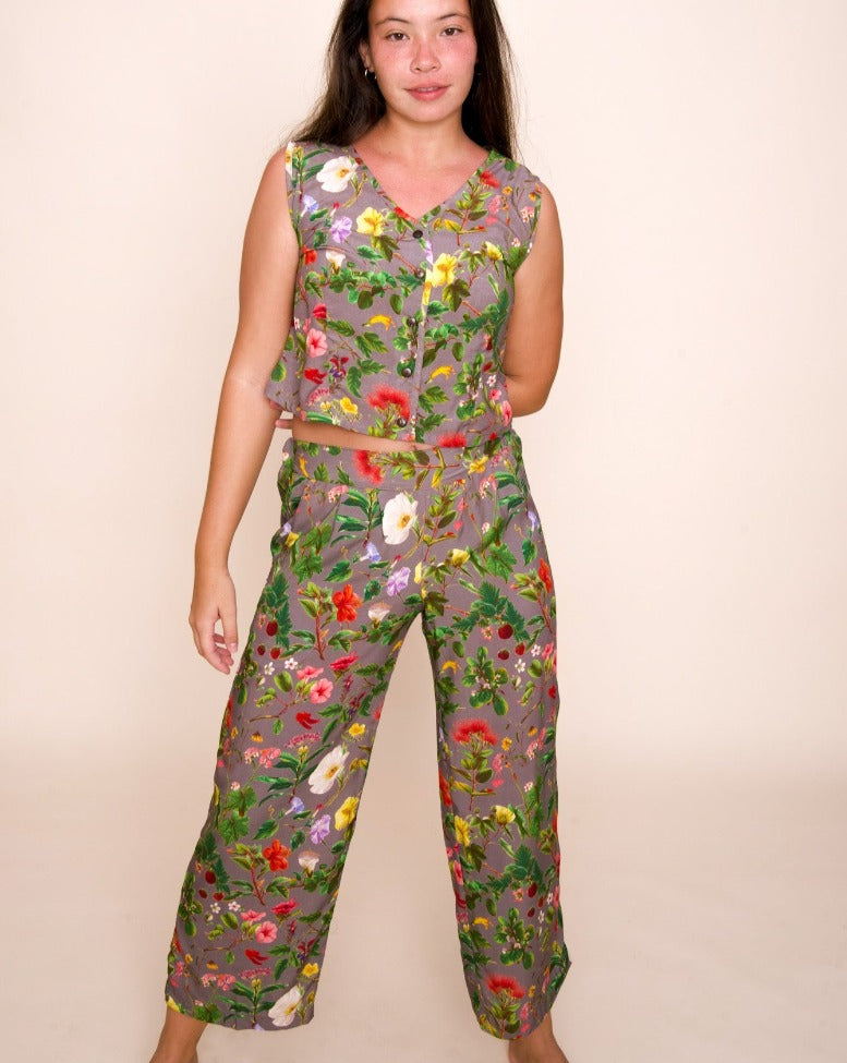 A model with long brown hair looks just beyond the camera, smiling slightly, while wearing a vest and pants with a complex pattern of native hawaiian plants on a grayish purple background.