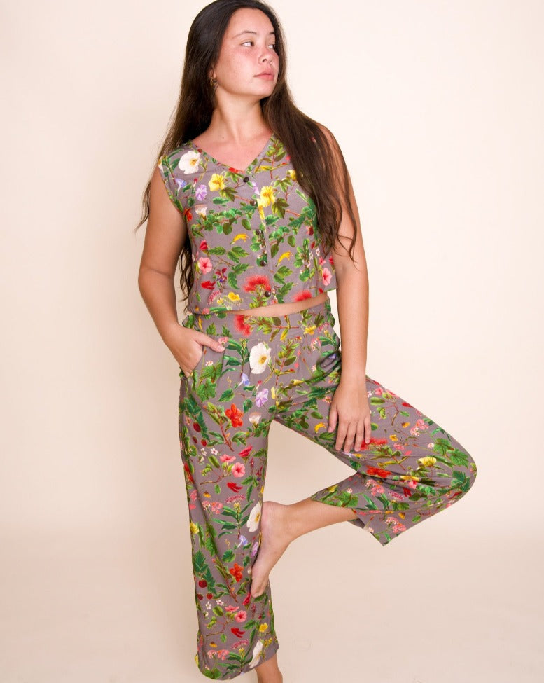 A model with long brown hair stands on one foot, with the other foot propped up on her knee. She looks to the left, and is wearing a cropped vest and pants that both feature a complex print of native hawaiian flowers.