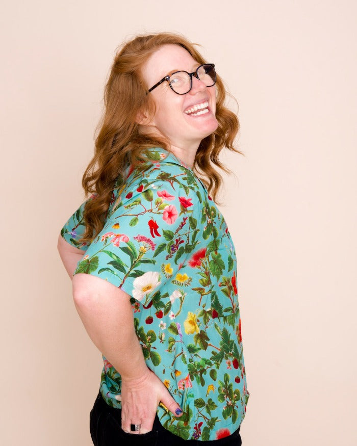 A model with medium-length red hair and glasses looks over their right shoulder, smiling at the camera, while wearing a teal colored shirt with an intricate pattern of native Hawaiian plants and flowers.
