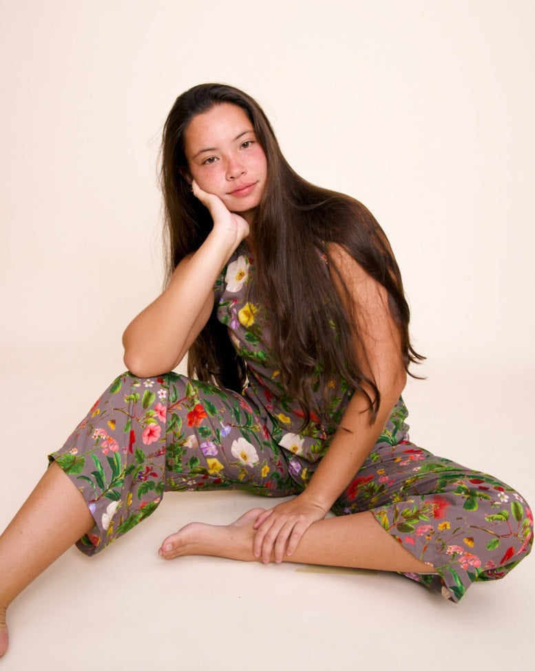 A model with long brown hair smiles into the camera with her cheek cupped in one hand. She sits on the floor and is wearing pants and a cropped vest with a complex pattern of native hawaiian pants on a grayist purple background.