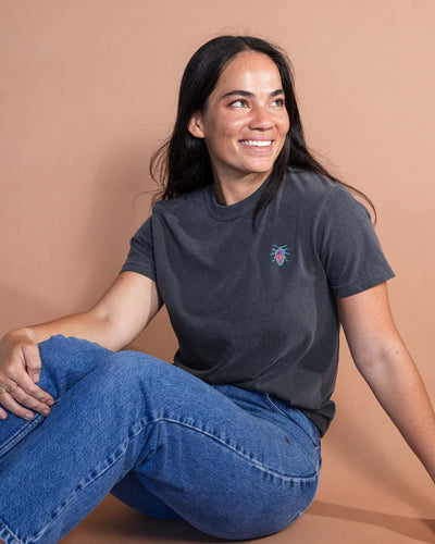 Woman sitting on the ground wearing jeans and a dark gray t-shirt with a small teal and red bug embroidered in the corner