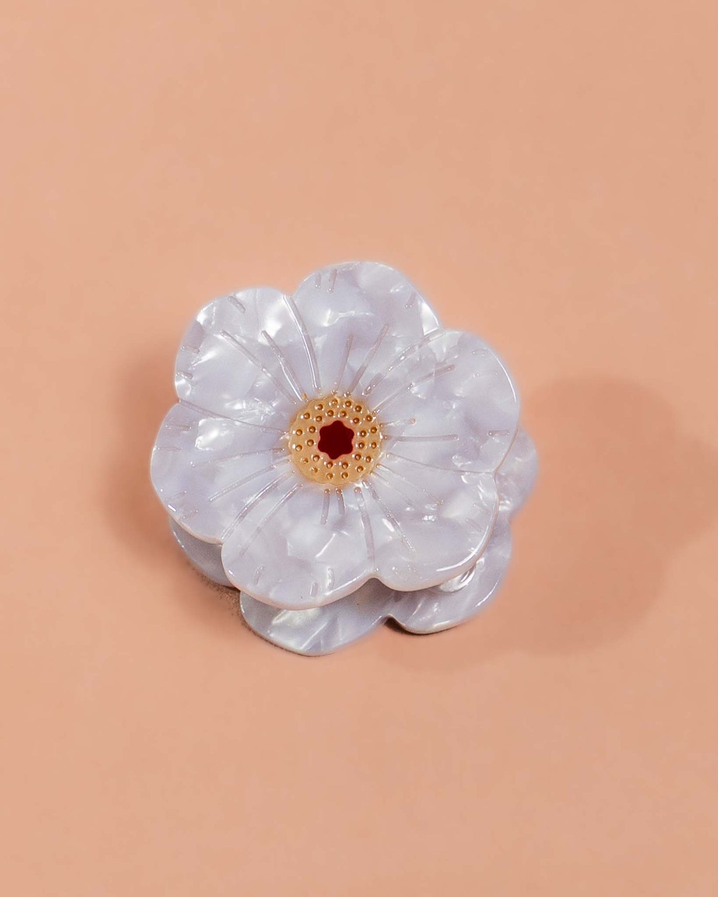A small hair clip of a flower with pearlescent white petals and a yellow center sits on a beige background