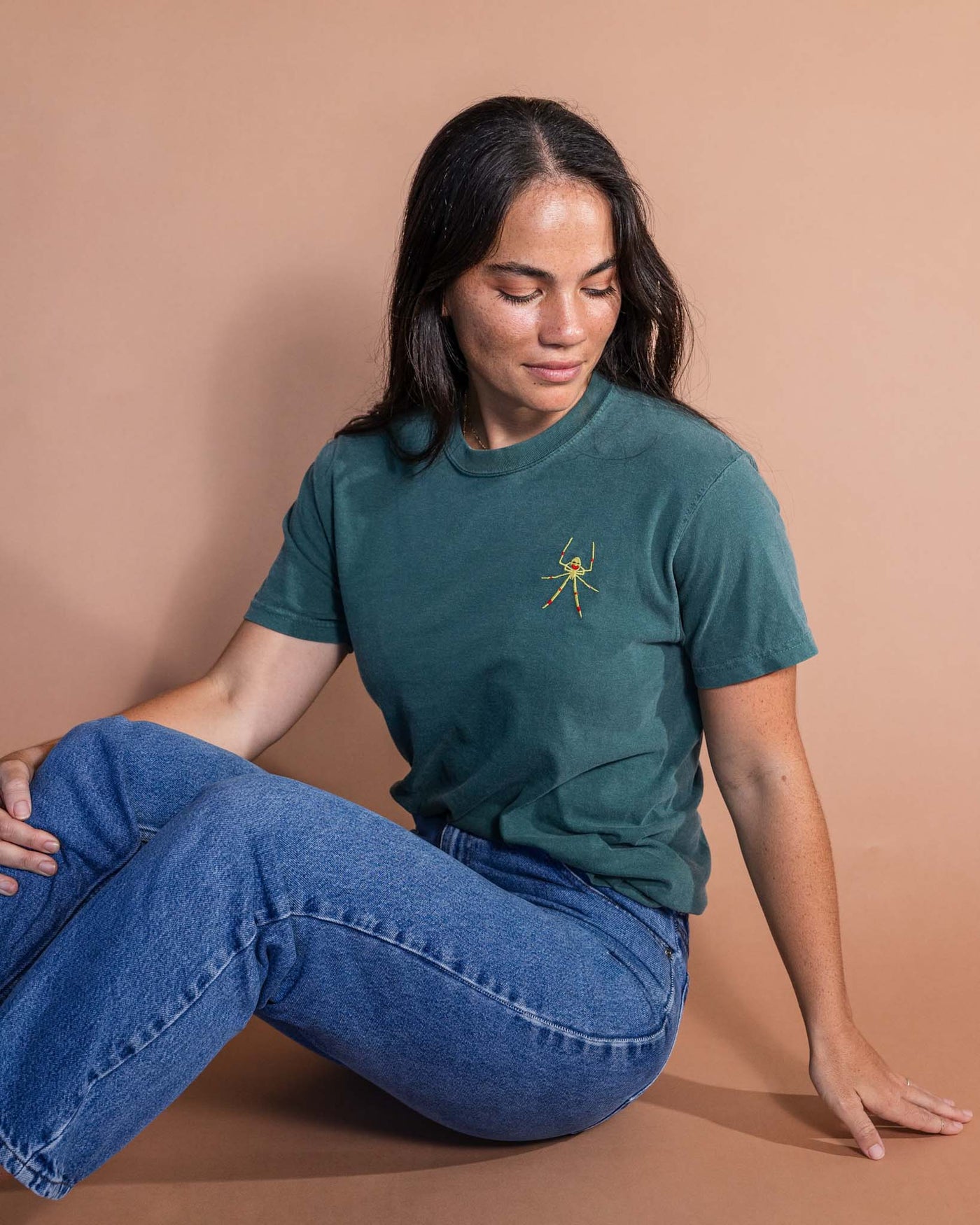 Woman wearing a teal t-shirt with a small yellow embroidered spider and blue jeans sitting on the ground