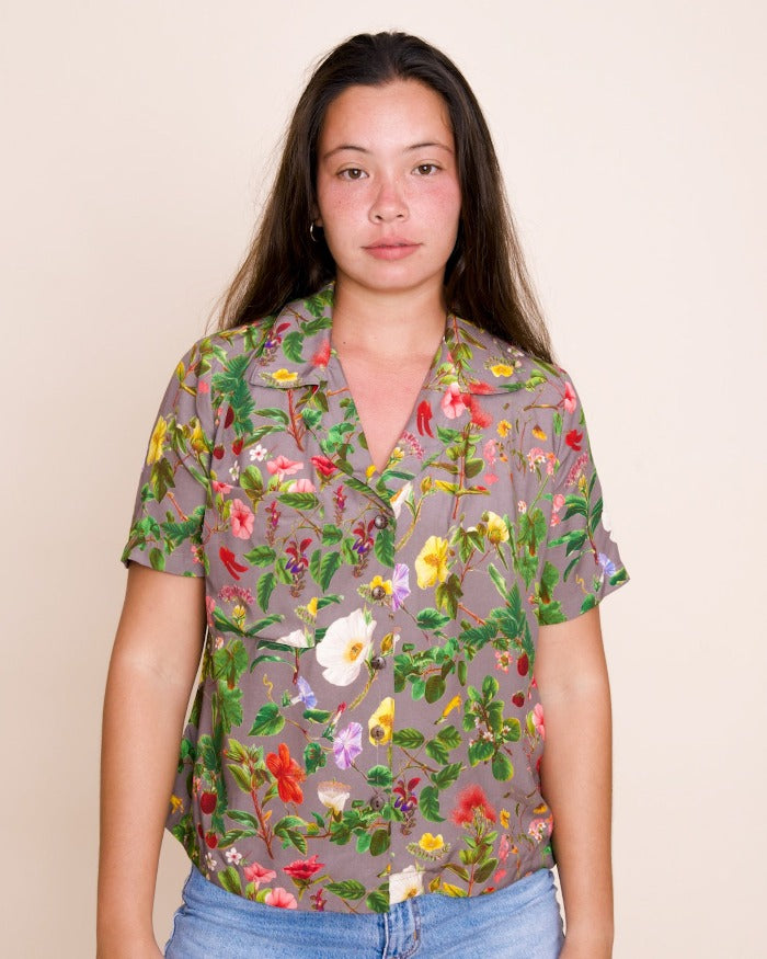 A model with long brown hair looks directly at the camera, wearing a poi-colored shirt with a delicate overall floral print.