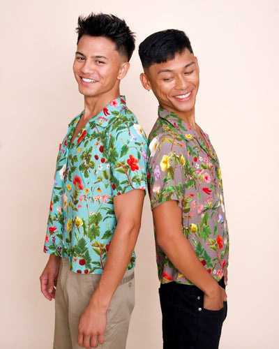 Two smiling models showcase button up, collared shirts with an intricate print of native Hawaiian plants. The model on the left has short brown hair and a kai-colored shirt, while the model on the right has short brown hair and a poi-colored shirt.