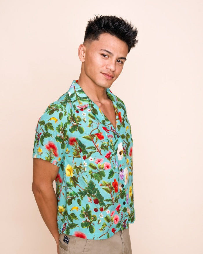 A model with short brown hair clasps his hands behind his back and looks just beyond the camera, smiling slightly and wearing a kai colored button up shirt with an intricate pattern of native Hawaiian plants and flowers.