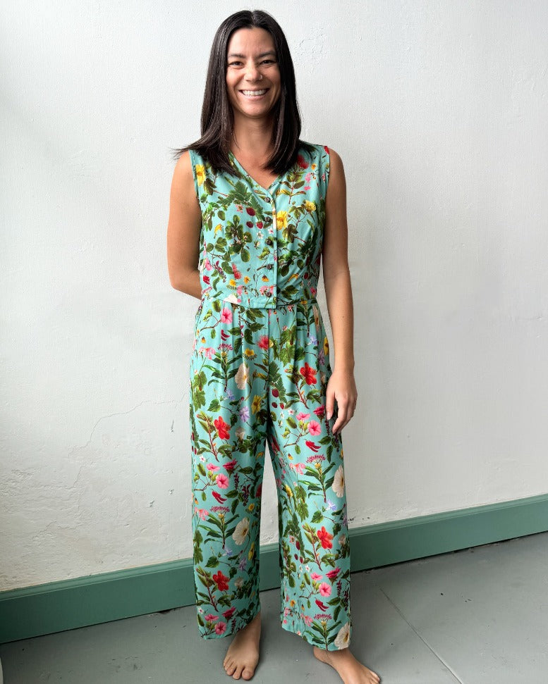 A model with medium length brown hair smiles happily at the camera, with her left arm at her side and her right arm behind her back. She's wearing a cropped vest and pants that are both a bright teal color with a complex pattern of native hawaiian flowers.