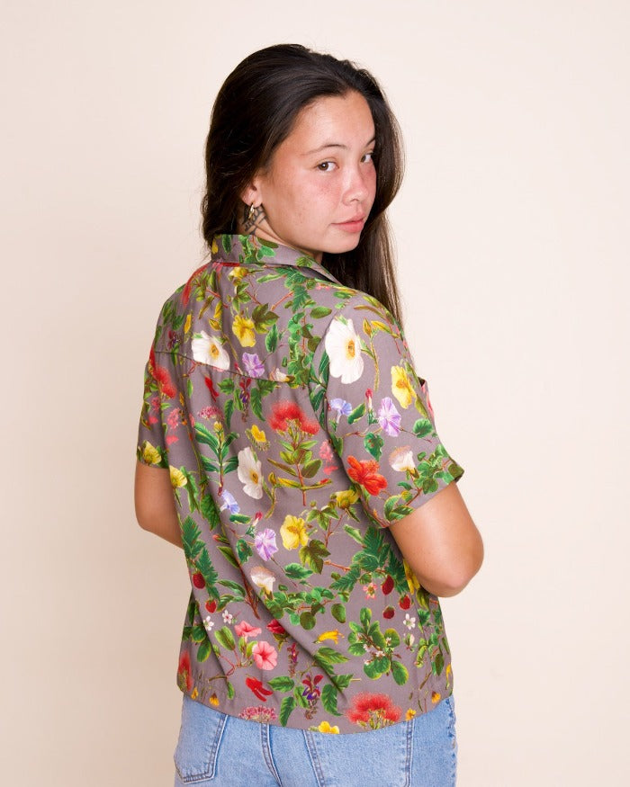 A model with long brown hair looks over their shoulder, wearing a poi-colored collared shirt with an intricate floral print.