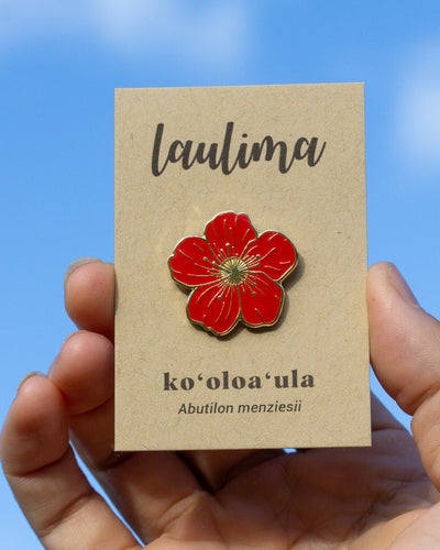 An enamel pin of a deep red flower with five broad petals is in the middle of a brown card that says "Laulima" at the top and "ko'oloa'ula" at the bottom.