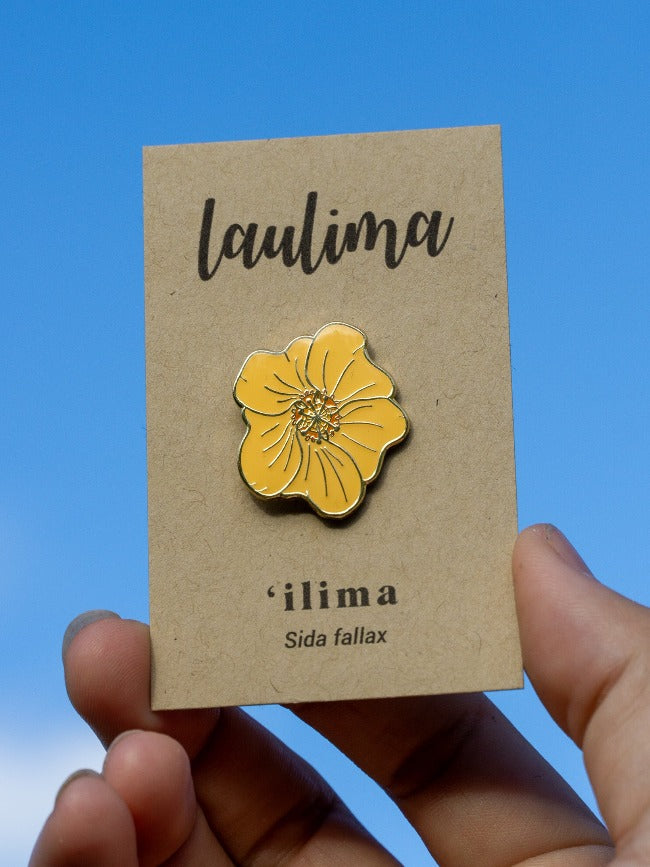 An enamel pin of a golden-yellow flower with five broad, overlapping petals is showcased on a brown paper card that says "Laulima" on the top and "'ilima" on the bottom.