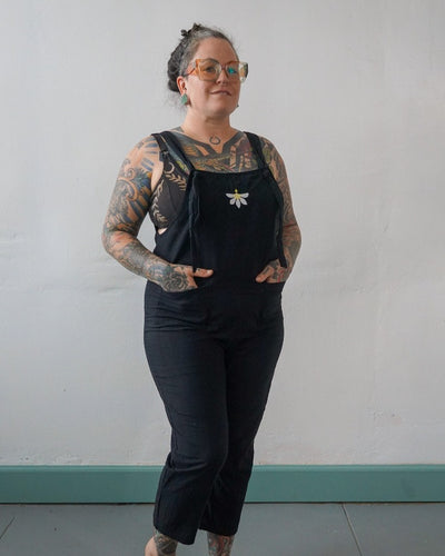 A heavily tattooed model with braided hair and glasses smiles confidently at the camera, with their hands in the pockets of black overalls with a white and yellow naupaka flower embroidered on the chest.