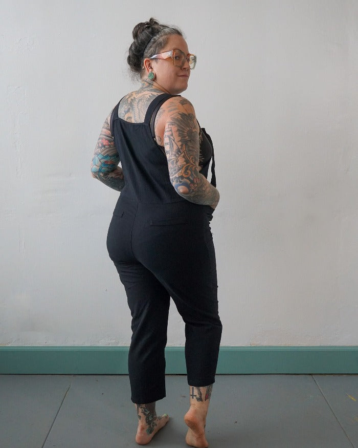 A model with full sleeve tattoos and glasses looks confidently over their shoulder, showcasing black linen overalls.