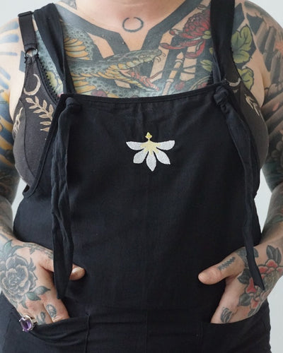 A white and yellow naupaka flower adorns the front of black, tie-front overalls.