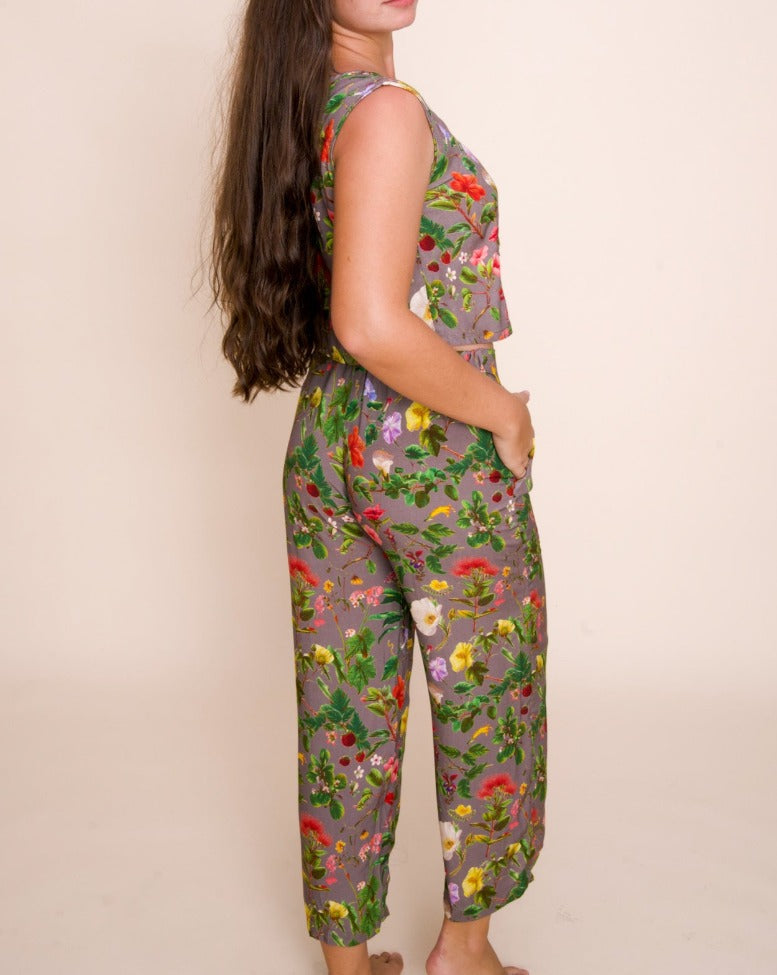 A model with long brown hair looks over her right shoulder with her right hand in her pocket. She's wearing pants and a cropped vest, both with a complex pattern on native hawaiian flowers.