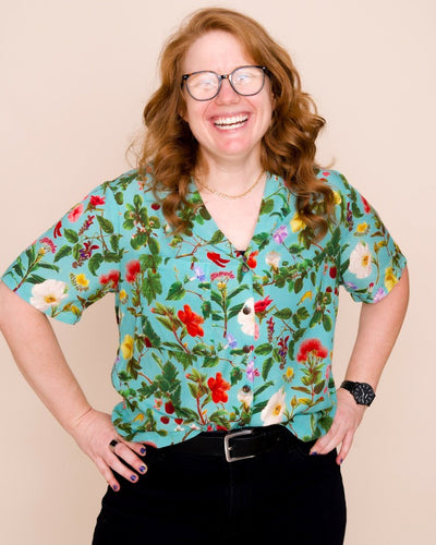 A model with medium-length red hair and glasses smiles at the camera, wearing a teal colored button up shirt with an intricate floral print of native Hawaiian plants.