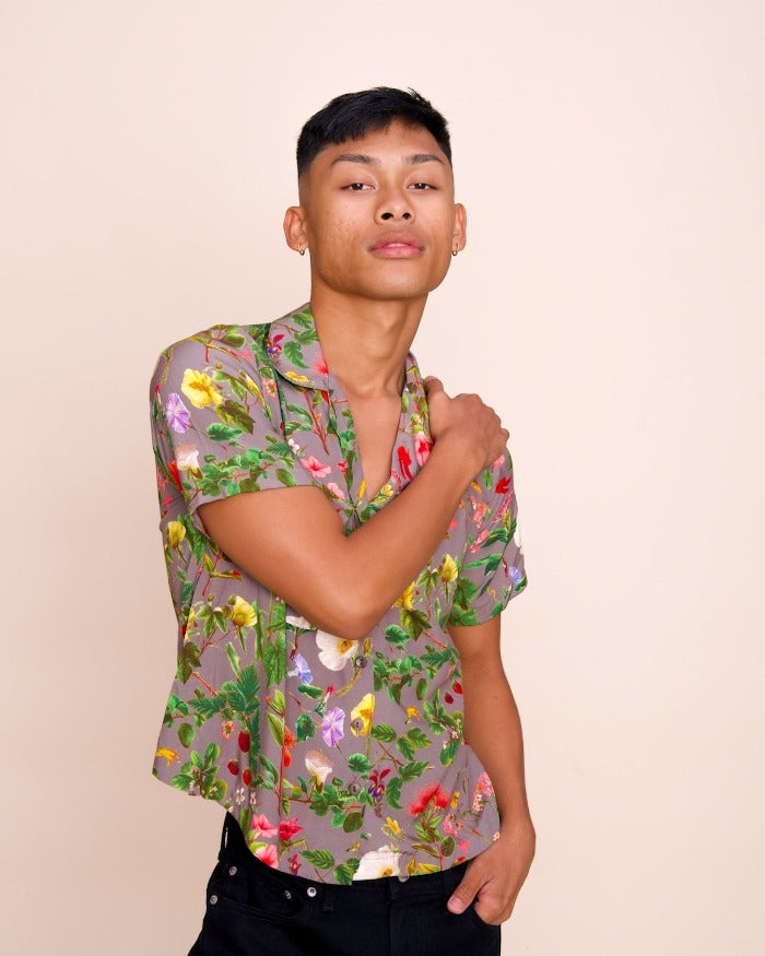 A model gazes confidently into the camera with their arm crossed over their chest, while wearing a poi-colored, button up shirt with an intricate floral pattern.
