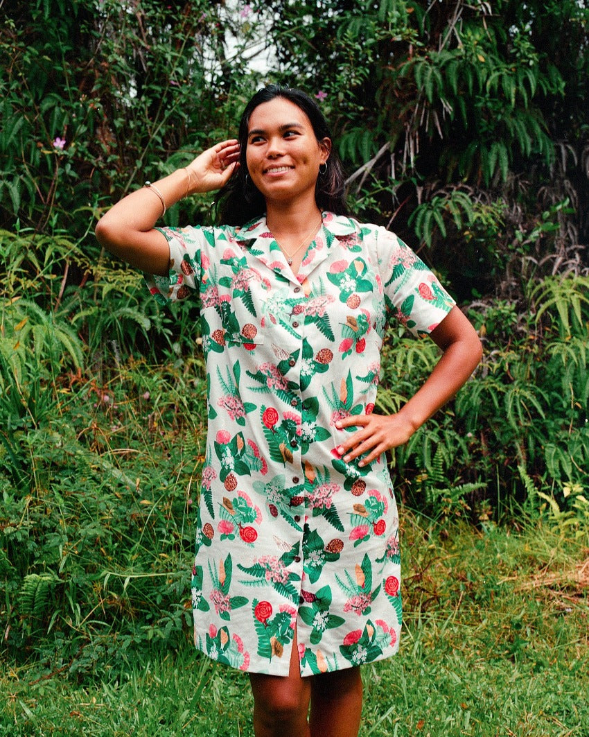 Kāhuli linen dress featuring Hawaiian land snails. Celebrating year of the Kāhuli. The smiling model faces the camera, and has long dark hair. She stands in front of a backdrop of ferns and forest.