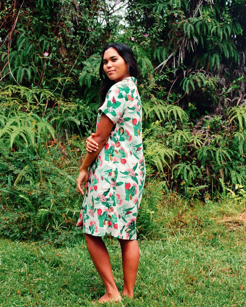Kāhuli linen dress featuring Hawaiian land snails. Celebrating year of the Kāhuli. The model has long dark hair and is standing barefoot in thick grass, in front of a backdrop of ferns and forest.